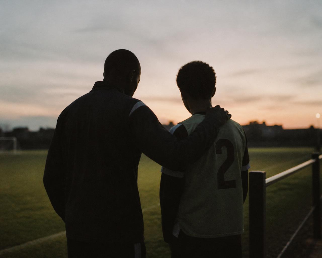 Treinador conversando com um jovem atleta após o treino, em silhueta contra o pôr do sol.